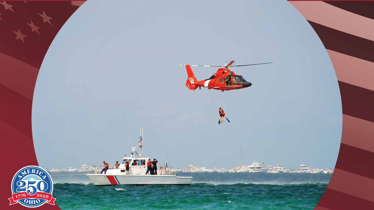 A Coast Guard helicopter flies above the ocean while a Coast Guard boat patrols the water below