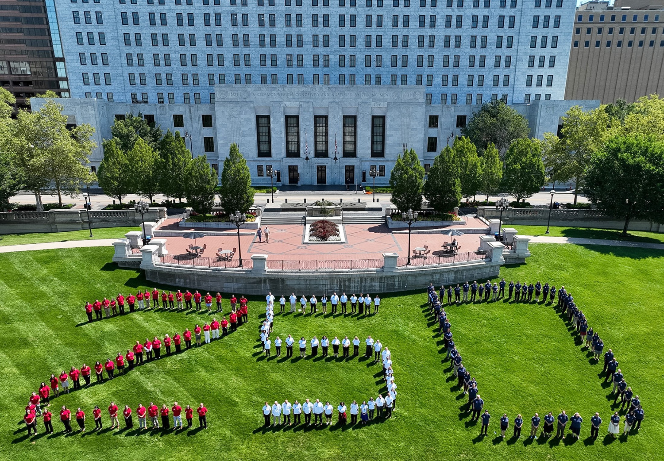 An aerial, wide-angle photograph captures a large group of people gathered on a vast green lawn in front of a massive, classical white stone building with rows of symmetric windows. The people are intentionally positioned to form the large number 