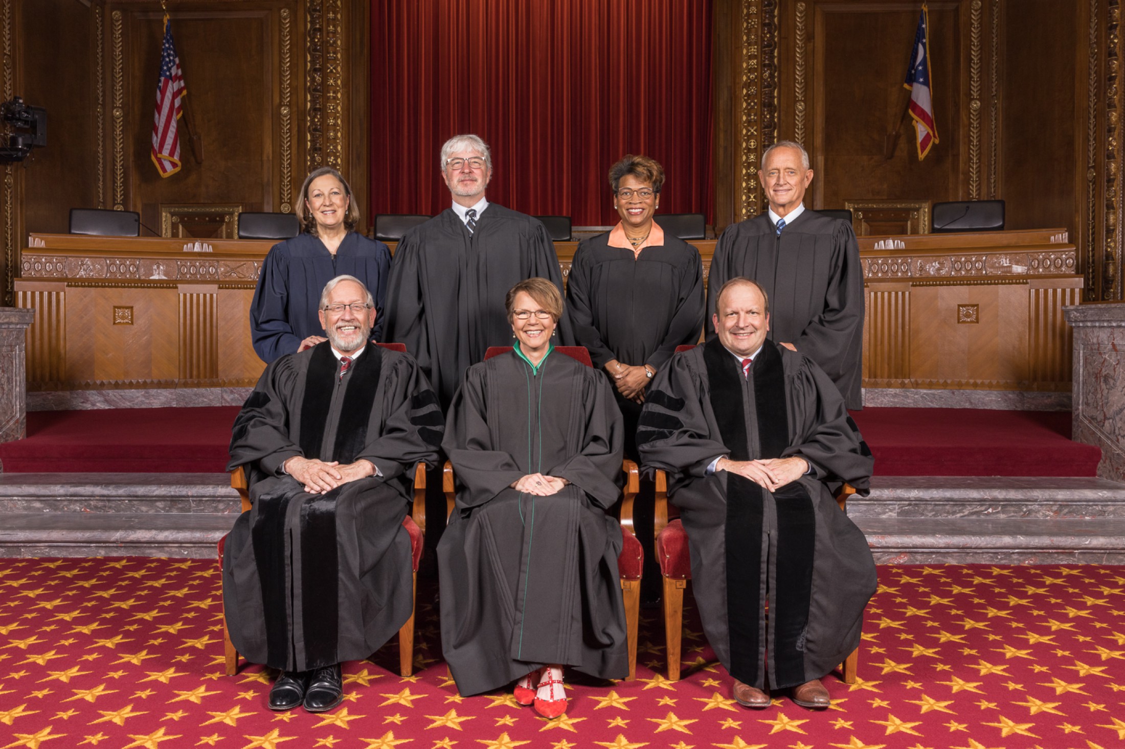 Image of seven men and women, all wearing black judicial robes. Two men and two women are standing and two men and one woman are seated in front of them in the courtroom of the Thomas J. Moyer Ohio Judicial Center.