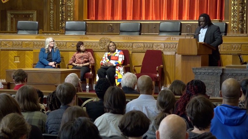 Image of three women sitting in red velvet chairs while a man stands at a wooden podium speaking to an audience of men and women in the courtroom of the Thomas J. Moyer Ohio Judicial Center.