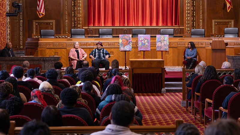 Image of a man and a woman sitting to one side of a display of three paintings. Another woman sits on the other side of the display. All are sitting in front of an audience of men and woman in the courtroom of the Thomas J. Moyer Ohio Judicial Center.