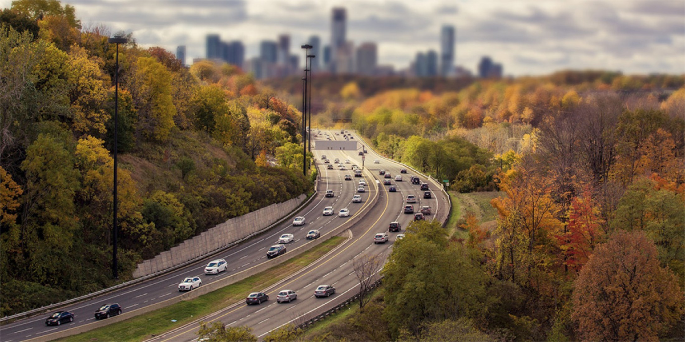 Image of a busy interstate, flanked on either side by fall foliage, and a city skyline in the background.