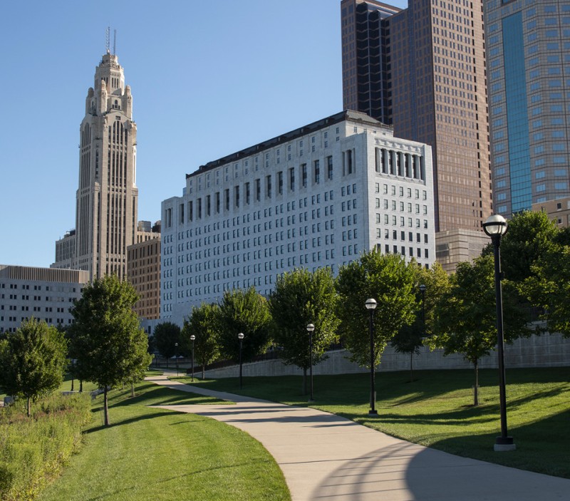 Image of a marble building with a brown roof sitting behind a walking path and several trees.