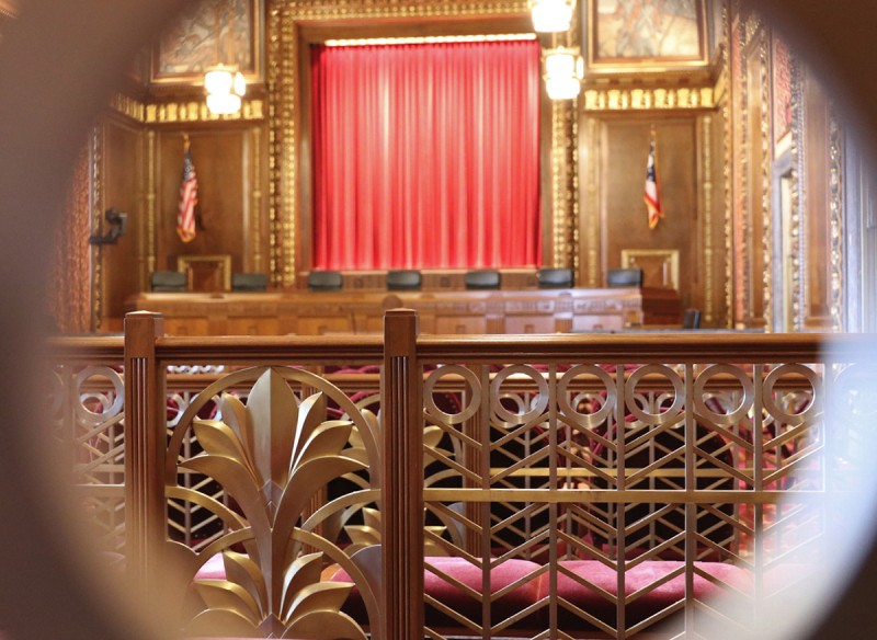 Image of an empty courtroom in the Thomas J. Moyer Ohio Judicial Center as seen through one of the ornate, bronze rings below the wooden handrails located throughout the courtroom.